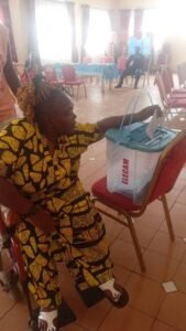 a woman is  seated in a red wheelchair. She reaches out with her right hand to deposit a paper ballot into a clear plastic ballot box labeled ELECAM.