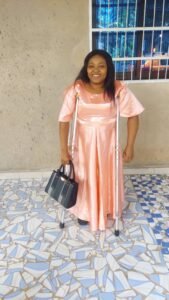 Armelle Nogning standing and smiling while using two underarm crutches. She is wearing a long, shiny peach-colored satin dress and holding a black handbag. She stands on a patio with blue and white mosaic tiling in front of a beige wall with a barred window.