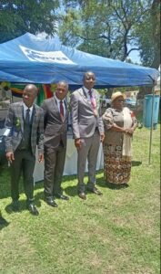 A full-length photo outdoors shows four people standing side-by-side beneath a blue tent with a white logo. From left to right: Professor Kudakwashe Tarubereka, wearing a grey plaid suit; Gift Mambipiri, in a dark suit with a burgundy tie; Councillor Nigel Rozario, wearing a light grey suit and a ceremonial collar/chain; and Marian Chombo, wearing a long patterned dress, a light shawl, and a straw hat. They are standing on a grassy area.