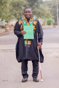 Kesah Princely stands outdoor after PhD Defense,  smiling as he holds a green thesis document and a white cane. 