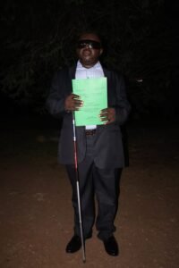 A full-length portrait of Ngong Peter Tonain standing in front of the Faculty of Social and Management Sciences, University of Buea, , holding his green PhD thesis and his white cane, wearing a black suit and dark glasses.