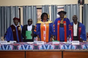 Ngong Peter Tonain, a visually impaired man in a suit holding his green thesis, stands with a four-member academic panel in blue and red regalia at the University of Buea after his PhD defense.