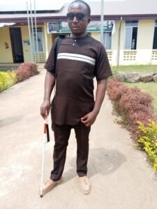 Prof. Charley R. Nugap, a visually impaired academic, stands on a walkway, wearing  a brown tunic set and sunglasses.