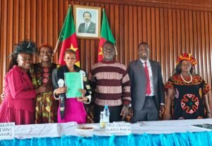 Six people stand behind a presentation table with handwritten name placards. Adele Nguizana stands center-left holding up a green folder, flanked by Dr Paule Marie Kougane, panel chair Prof. Blasius Chiatoh, and other academic officials, in front of a wooden wall with Cameroonian flags.