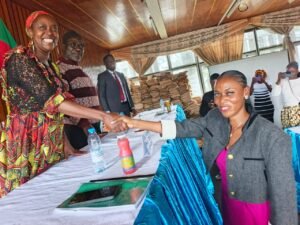 Adele Nguizana, wearing a dark jacket over a pink dress, smiles as she shakes hands across a conference table with her supervisor, Dr Paule Marie Kougane, who wears a colorful red-patterned dress. A green thesis folder rests on the table between them.