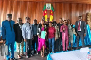 Adele Nguizana stands at the center of a group of supporters and academics inside a Faculty of Arts' Boardroom, holding a green dissertation folder and a white cane. Several visually impaired participants alongside her hold white canes, with Cameroonian flags and a portrait of President Paul Biya displayed on the wall behind them.