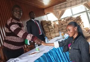 Adele Nguizana reaches across a draped table to shake hands with panel chairperson Prof. Blasius Chiatoh, who wears a brown pattern sweater and glasses. Placards and refreshments are visible on the table.