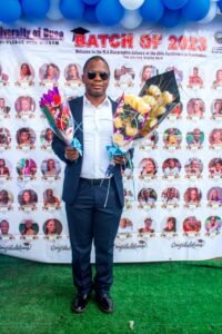 A full-length photograph of visually impaired researcher Mr. Ngifor Ronald standing outdoors on green artificial turf. He is wearing a dark blue suit, white shirt, and dark sunglasses while holding two celebratory flower bouquets. Behind him is a large University of Buea graduation banner lined with blue and white balloons.