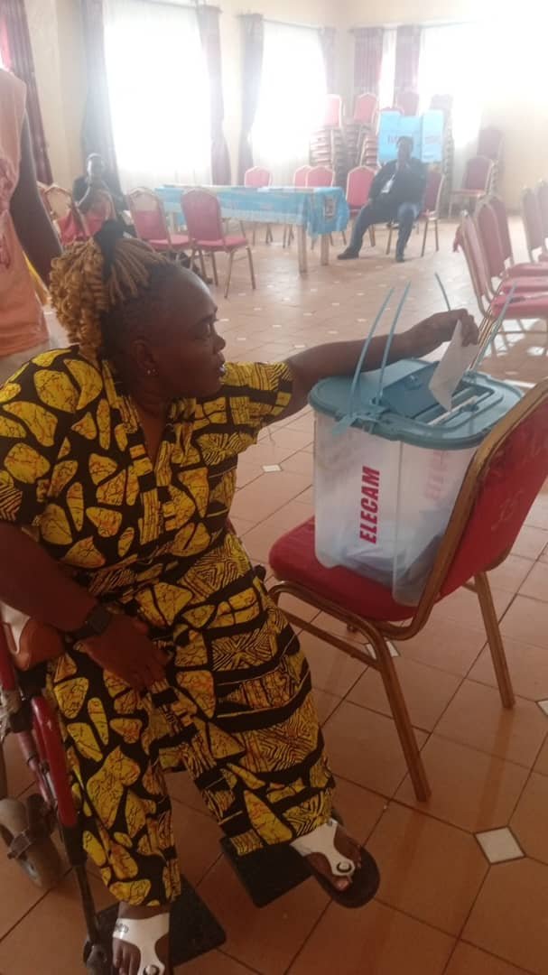 a woman is seated in a red wheelchair. She reaches out with her right hand to deposit a paper ballot into a clear plastic ballot box labeled ELECAM.