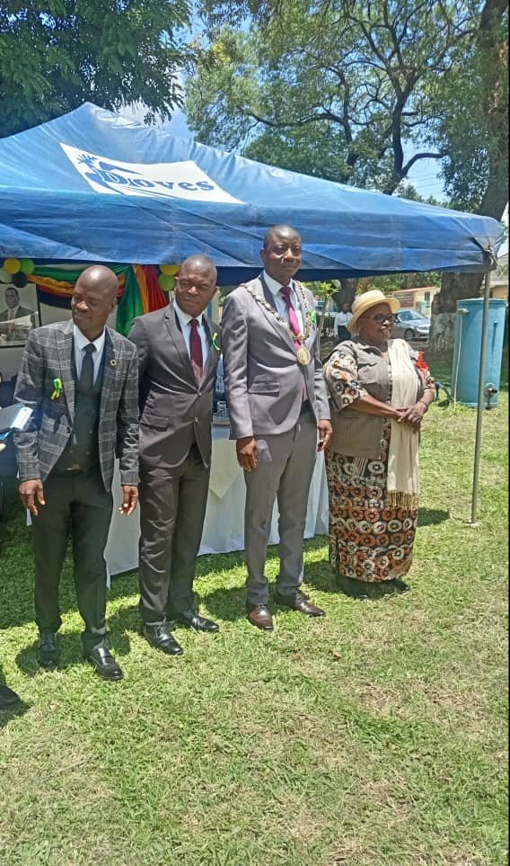 A full-length photo outdoors shows four people standing side-by-side beneath a blue tent with a white logo. From left to right: Professor Kudakwashe Tarubereka, wearing a grey plaid suit; Gift Mambipiri, in a dark suit with a burgundy tie; Councillor Nigel Rozario, wearing a light grey suit and a ceremonial collar/chain; and Marian Chombo, wearing a long patterned dress, a light shawl, and a straw hat. They are standing on a grassy area.