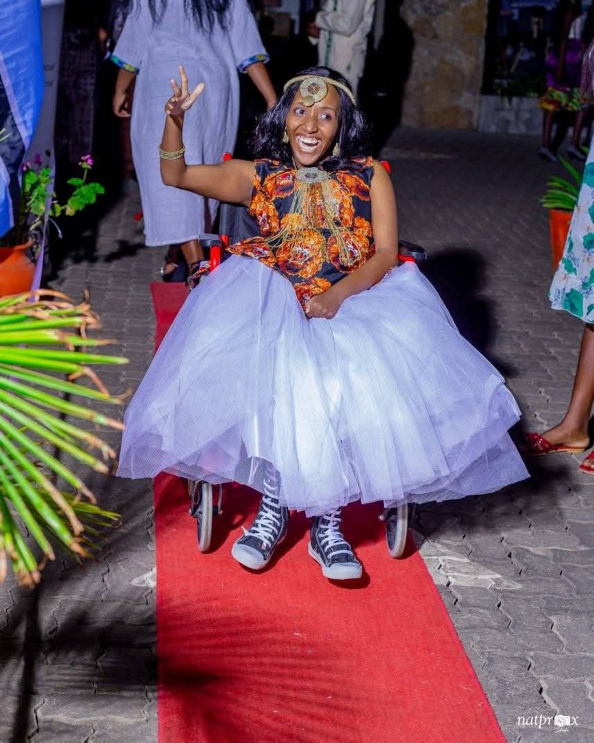 Esther Munetsi, a model who is rewriting beauty standards, smiles joyfully as she makes a grand entrance on a red carpet in her wheelchair. She wears a vibrant orange and black patterned bodice paired with a voluminous white tulle skirt and black high-top sneakers. Her look is completed with a gold headband and jewelry, and she holds up a peace sign as she moves forward.