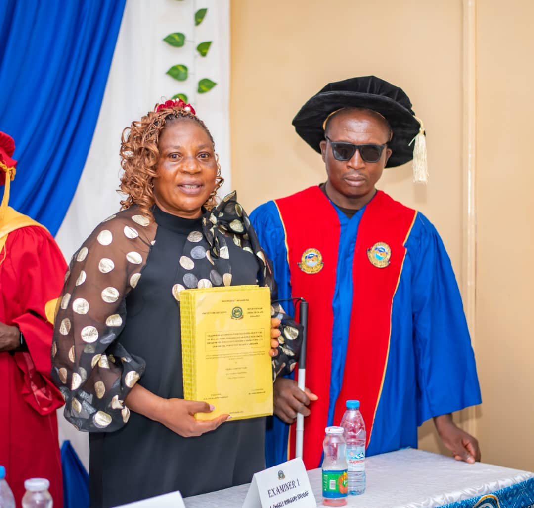 A medium shot at the University of Bamenda showing PhD candidate Comfort Yigha and Professor Charley R. Nugap. Comfort, on the left, wears a black blouse with sheer polka-dot sleeves and holds a thick, yellow-bound thesis. To her right, Prof. Nugap, who is visually impaired, wears a blue and red academic gown, a black doctoral hat, and sunglasses while holding a white cane. They stand behind a table.