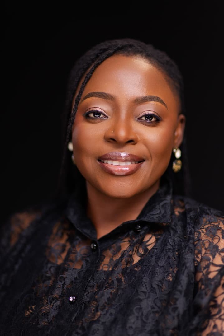 Comfort Mussa, author of The Shape of Unbroken Things/Ce Qu'on Ne Brise Pas, smiles warmly against a plain black background. She has dark braided hair, framed makeup, and is wearing a black lace collared blouse with drop earrings.