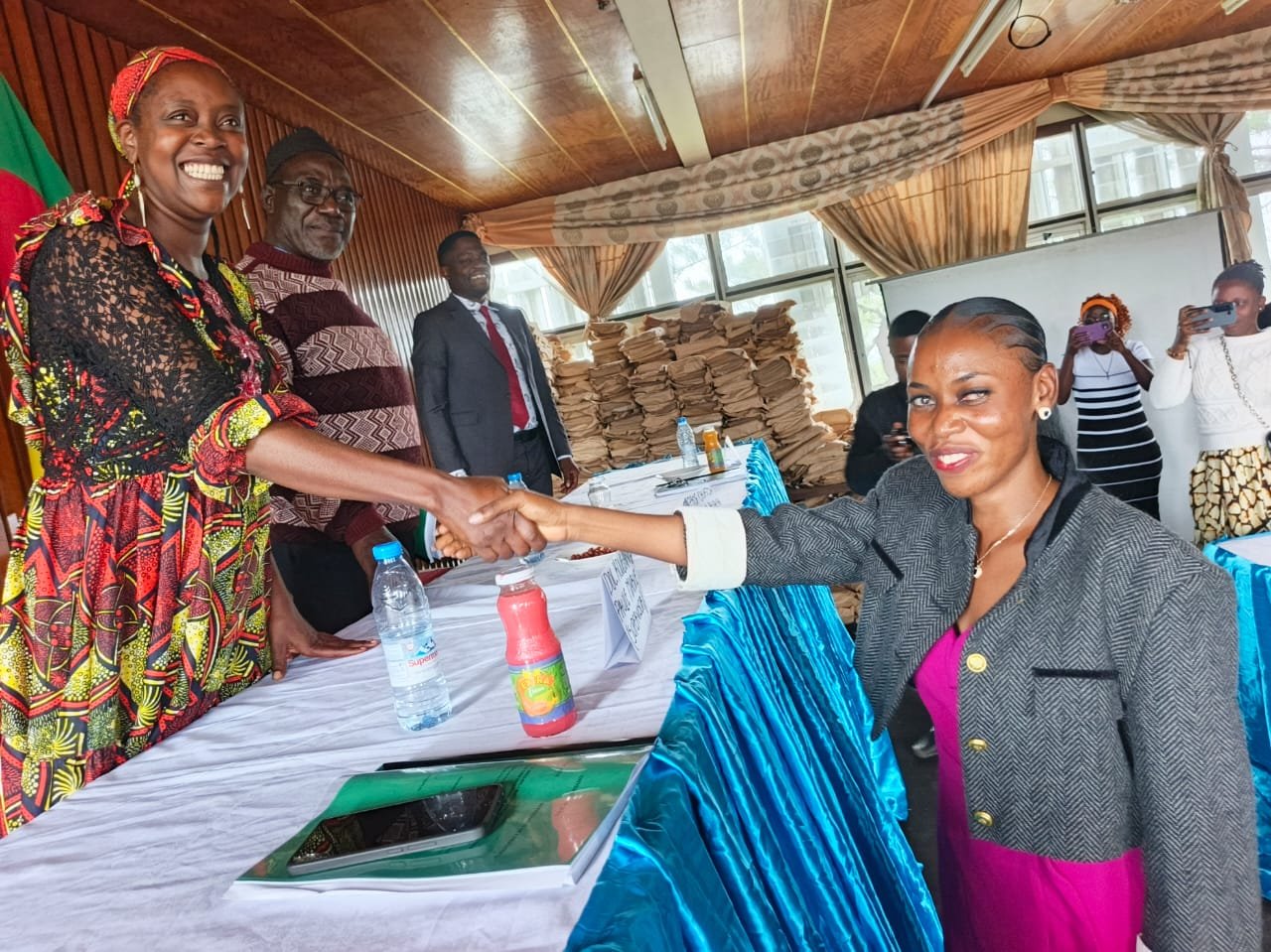 Adele Nguizana, wearing a dark jacket over a pink dress, smiles as she shakes hands across a conference table with her supervisor, Dr Paule Marie Kougane, who wears a colorful red-patterned dress. A green thesis folder rests on the table between them.