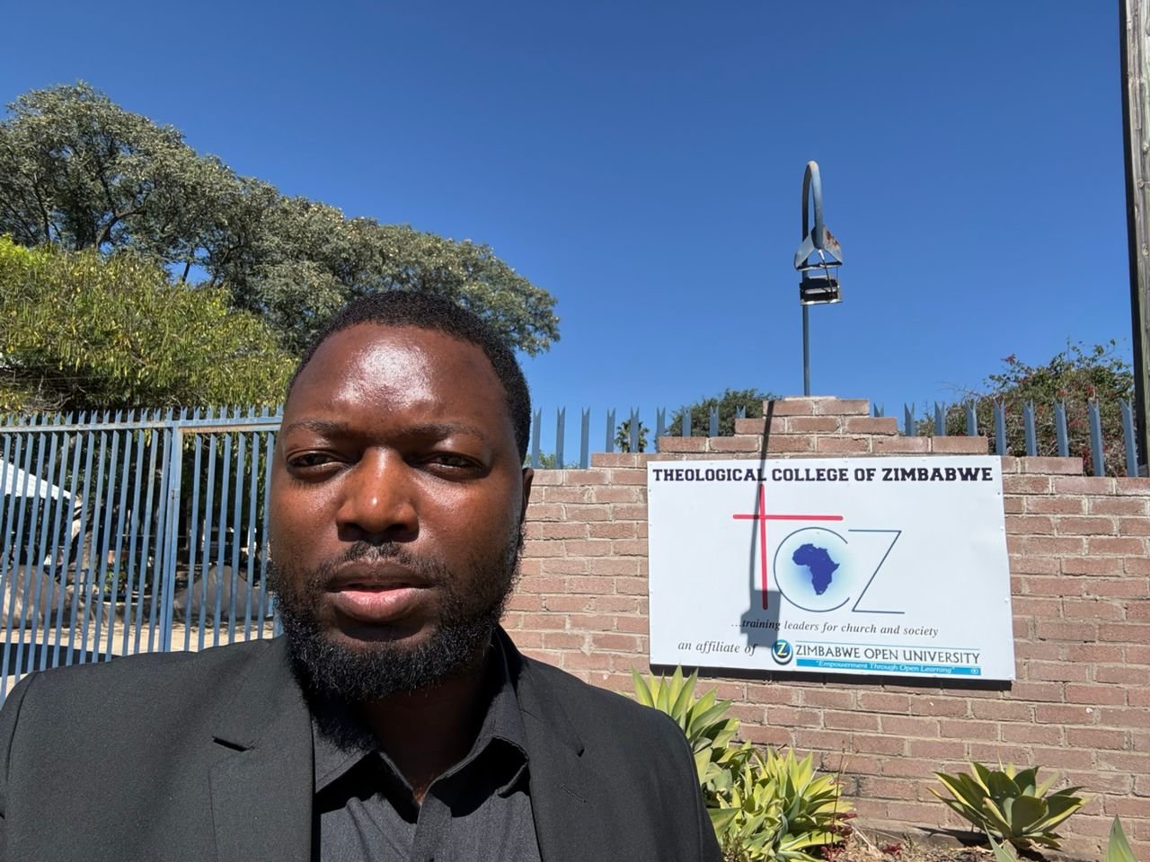 An outdoor portrait of Precious Madzimbe, a Black man with a dark beard and short dark hair, wearing a black suit jacket and dark shirt. He is standing under a clear blue sky in front of a brick wall and metal fence. Mounted on the brick wall is a white sign that reads "THEOLOGICAL COLLEGE OF ZIMBABWE ...training leaders for church and society an affiliate of ZIMBABWE OPEN UNIVERSITY".