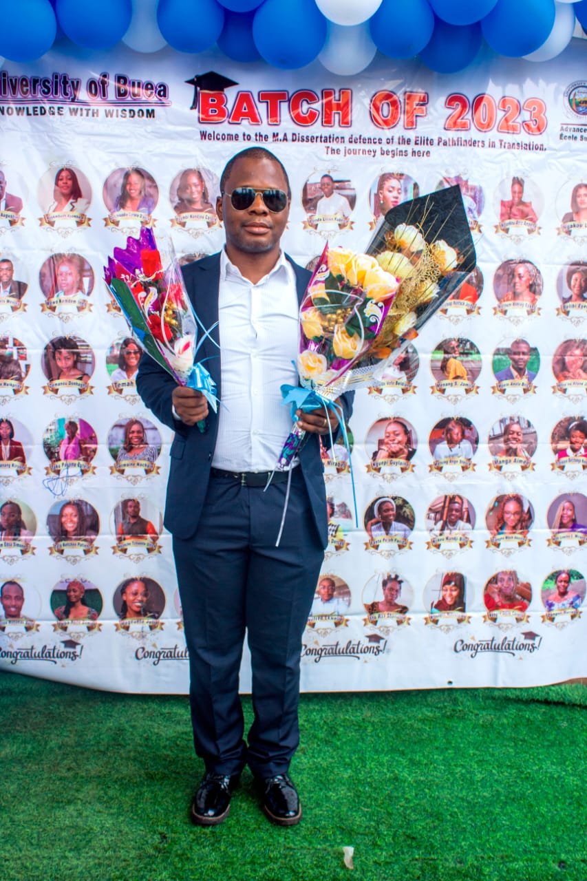 A full-length photograph of visually impaired researcher Mr. Ngifor Ronald standing outdoors on green artificial turf. He is wearing a dark blue suit, white shirt, and dark sunglasses while holding two celebratory flower bouquets. Behind him is a large University of Buea graduation banner lined with blue and white balloons.